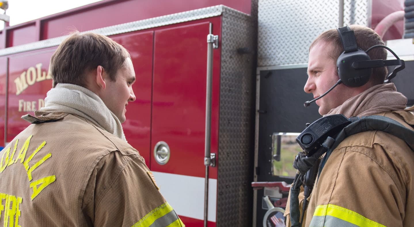 Firefighters wearing FIRECOM headsets beside a red engine, talking through an apparatus intercom