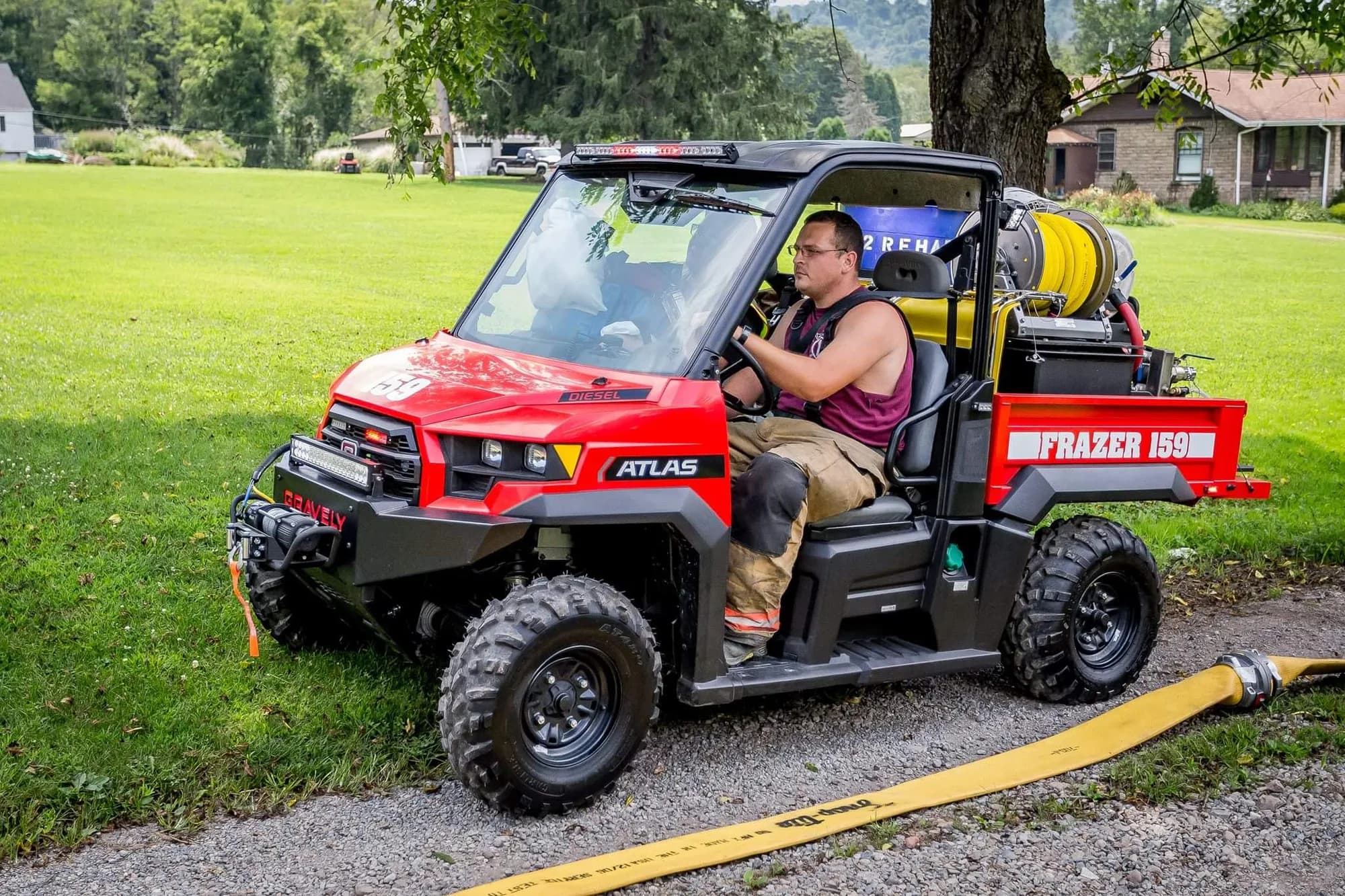 Firefighter driving a red Gravely Atlas JSV diesel side-by-side lettered Frazer 159, carrying a bed-mounted fire skid with a hose reel, tank, and plumbed pump module, with a supply line laid out on the gravel behind it