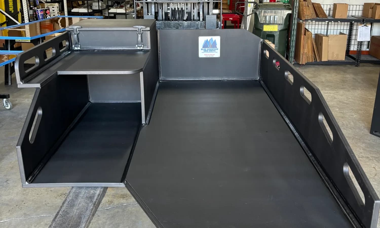 Bare Blue Mountain medical skid body on the shop floor: an open flat patient deck on the right, a raised shelf with a latching lidded compartment and an open storage bay on the left, low slotted side rails, and a serial-numbered Blue Mountain Skid Units plate on the headboard.